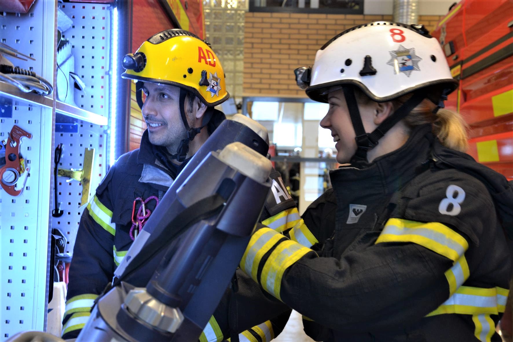 En man och en kvinna iklädda räddningstjänstens utryckningsuniform står vid en brandbil i ett garage.